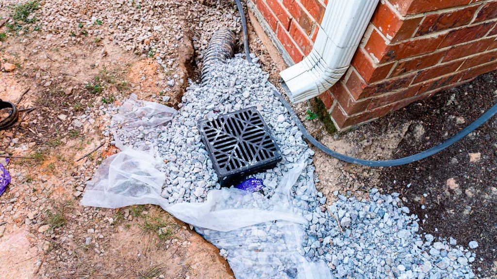 Square drainage grate in the ground with a crushed rock near a downspout